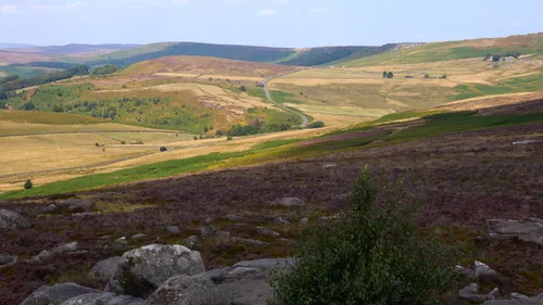 This landscape photograph captures the scenic beauty of Stanage Edge in the Peak District, England, during the early afternoon in late summer. The image showcases rolling hills carpeted with a vibrant mix of green and purple heather, characteristic of this season, with rocky outcrops in the foreground. The sweeping views reach towards the Longshaw Estate, set amidst patchwork fields and gently winding roads. Stanage Edge stands out as the main subject, a prominent gritstone escarpment ideal for walking and climbing, with its spectacular vantage over the heart of the United Kingdom’s countryside. The clear sky and warm sunlight enhance the natural colours, providing an idyllic representation of the Peak District and its picturesque summer landscape.